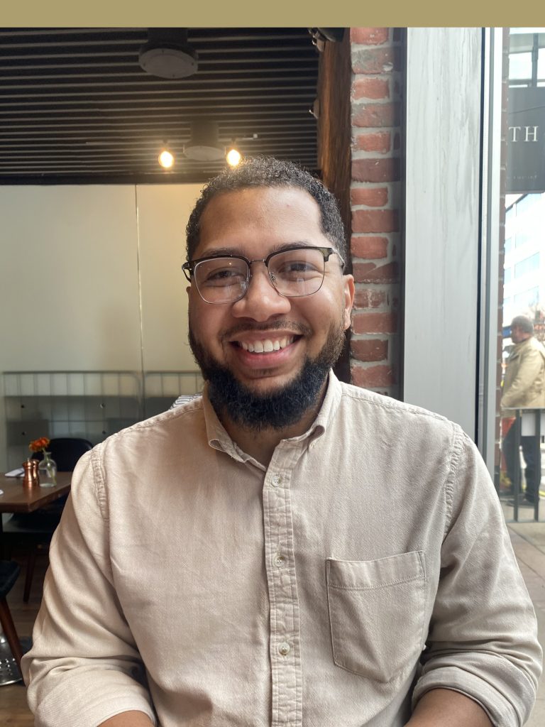 Headshot of Anthony Jones, Supervisor of the Year. He is wearing a brown button-up shirt and glasses, sitting in a brick-walled cafe.