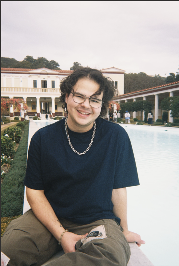 Headshot of Skyler Kasnoff, Undergraduate Student Employee of the Year. He is wearing a black tshirt and a chain necklace, sitting next to a large pool